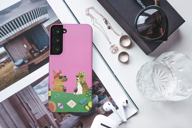 May include: A pink phone case with a cartoon illustration of two dogs wearing sunglasses and drinking wine at a poker table. The image also includes sunglasses, jewellery, rings, a glass, and wireless earbuds.