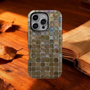 Puede incluir: Una funda para smartphone con un diseño de mosaico en tonos marrones, verdes y azules. La funda tiene un acabado brillante y un recorte para la cámara. La funda del teléfono está sobre una superficie de madera con un libro en el fondo.