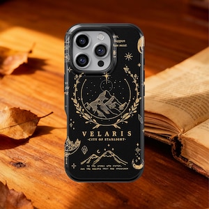 Puede incluir: Funda de teléfono negra con un diseño dorado de montaña y estrellas. La funda presenta el texto "VELARIS - CITY OF STARLIGHT" y otras inscripciones. La funda está sobre una superficie de madera con un libro.