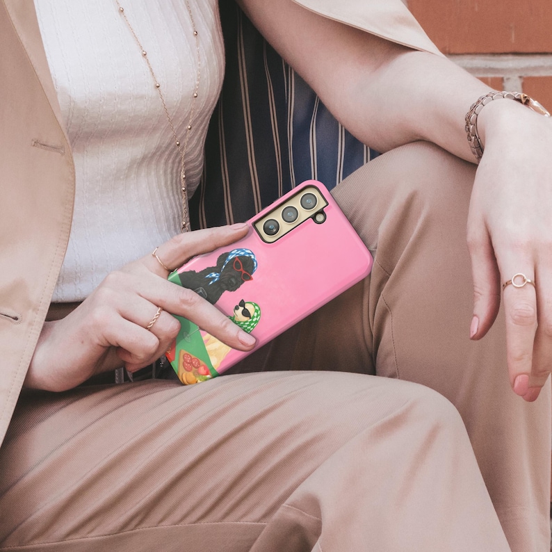 May include: A pink smartphone case with a vibrant graphic of two figures wearing sunglasses. The case has cutouts for the camera and buttons. The phone case is held by a person wearing rings and a watch.