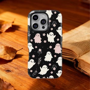 Puede incluir: Funda de teléfono negra con un patrón repetido de fantasmas blancos y rosas, pequeñas flores rosas y ramitas verdes. La funda tiene un acabado brillante y un borde negro. La funda del teléfono está sobre una superficie de madera.