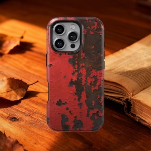 May include: A smartphone case with a distressed red and brown paint effect. The case has a black border and a camera cutout. The background is a wooden surface with a book and leaves.