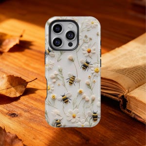 May include: White phone case featuring a 3D floral and bee design. The case has a raised texture with white daisies, green stems, and yellow and black bees. The phone case is on a wooden surface.
