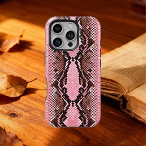 Może przedstawiać: Różowa i brązowa obudowa telefonu z wzorem skóry węża. Obudowa ma teksturowany wzór i wycięcie na aparat. Obudowa leży na drewnianej powierzchni z książką i liśćmi w tle.