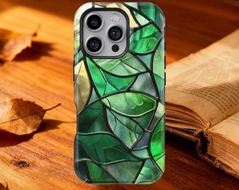 May include: A smartphone case with a stained-glass leaf design in shades of green and yellow. The case has a black border and a camera cutout. The case is on a wooden surface with a book and dried leaves.
