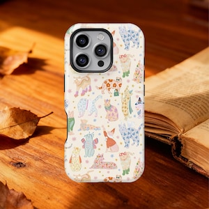 Puede incluir: Una funda de teléfono con un diseño lúdico de gatos ilustrados en diferentes poses y atuendos. Los gatos son en tonos de rosa, azul, naranja y verde, sobre un fondo crema con pequeños acentos de estrellas. La funda se muestra sobre una superficie de madera.