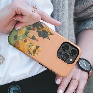 May include: A peach-coloured phone case with a quirky illustration of two cats drinking wine. One cat wears sunglasses and a scarf. The case has a black camera module and a green top edge.