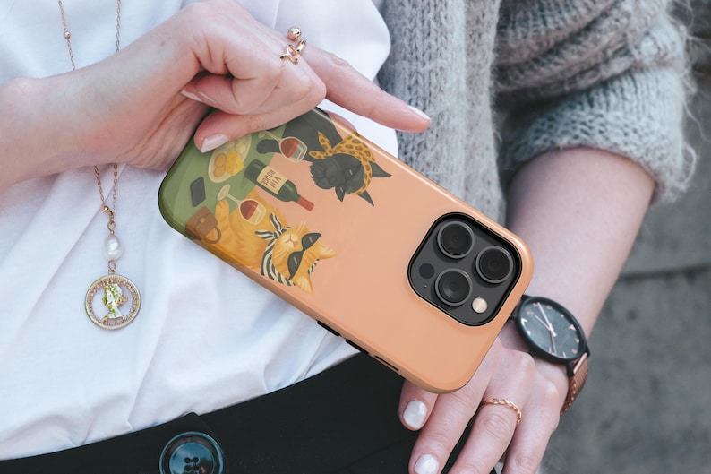 May include: A peach-coloured phone case with a quirky illustration of two cats drinking wine. One cat wears sunglasses and a scarf. The case has a black camera module and a green top edge.