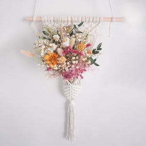 May include: A white macrame wall hanging featuring a wooden dowel and a woven pocket holding a bouquet of dried flowers. The arrangement includes white daisies, pink baby's breath, orange carnations, yellow billy balls, and green eucalyptus leaves.