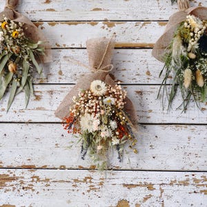 May include: Three bundles of dried flowers wrapped in natural tan hessian fabric. Each arrangement features a mix of white, cream, orange, and green dried botanical elements, including small daisies, baby's breath, and long thin green leaves.