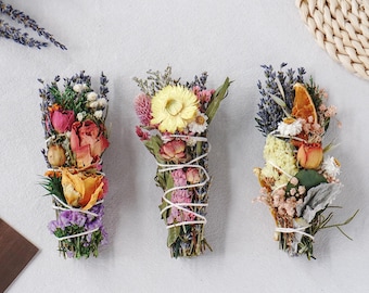 May include: Three bundles of dried flowers and herbs tied with white string. The left bundle features pink and orange roses with purple lavender. The center bundle has a large yellow daisy, pink clover, and greenery. The right bundle includes dried orange slices, yellow flowers, and lavender.