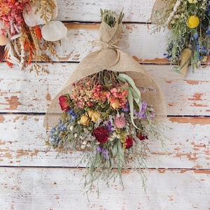 May include: A bouquet of dried flowers wrapped in tan hessian fabric. The arrangement features red, yellow, pink, purple, and blue blossoms mixed with green foliage and thin stems. The stems are tied together with a matching hessian twine string.