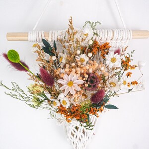 May include: A wall hanging featuring a macrame pocket suspended from a wooden dowel, filled with a dried floral arrangement. The bouquet includes white daisies, orange statice, green foliage, and deep purple fuzzy grass plumes.