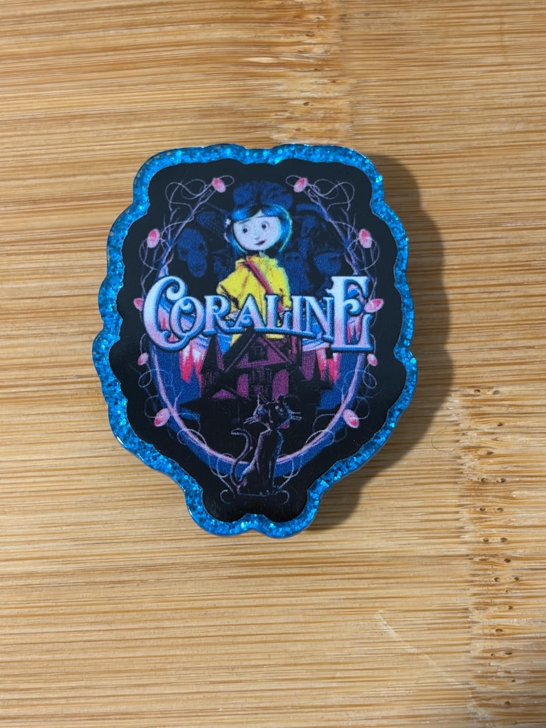 May include: A decorative pin featuring a blue glitter border and a central illustration of the character Coraline in a yellow raincoat, a black cat, and a house silhouette. The text Coraline is printed in a stylised font across the centre.