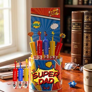 May include: A comic book-themed gift set with a "Super Dad" design. It includes a cylindrical container and a card with comic book graphics. Several pens with superhero-themed toppers are displayed, with text like "My real-life hero".