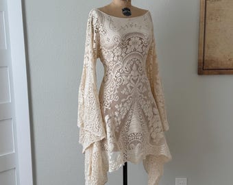May include: An off-white, cream-colored lace dress featuring a wide scoop neckline, long flared bell sleeves, and an asymmetrical hemline. The intricate floral and scrollwork lace pattern covers the entire garment, which is displayed on a mannequin.