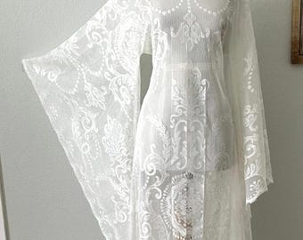 May include: A floor-length white sheer dress featuring an intricate embroidered floral pattern, a wide scoop neckline, and long, flowing bell sleeves. The garment is displayed on a mannequin, showcasing the detailed lace-like fabric texture.