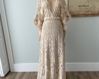 May include: A cream-colored, floor-length lace dress with a plunging neckline and long, bell sleeves. The dress is displayed on a mannequin and features intricate floral patterns. The sleeves have a fringed trim. The dress is a vintage or bohemian style.