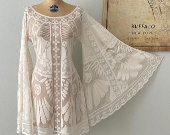 May include: A cream-colored lace dress featuring a wide scoop neckline, long flared bell sleeves, and an intricate floral pattern throughout. The garment is displayed on a beige dress form, showcasing a scalloped hem and detailed mesh texture.