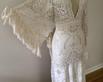 May include: A floor-length off-white dress featuring intricate lace patterns throughout. The garment has a deep V-neckline, wide kimono-style sleeves with long fringe trim along the edges, and a fitted bodice that transitions into a flowing skirt.