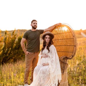review photo of an actual customer and her fiancé she is modeling a bohemian lace wedding dress with a crochet bodice and long bell sleeves that are lined with fringe. She is pregnant and sitting in a chair. This is a maternity photo shoot for her wedding day.
