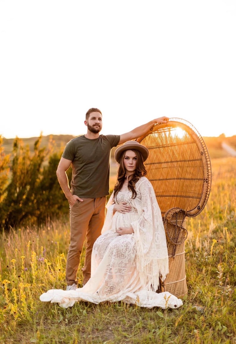 review photo of an actual customer and her fiancé she is modeling a bohemian lace wedding dress with a crochet bodice and long bell sleeves that are lined with fringe. She is pregnant and sitting in a chair. This is a maternity photo shoot for her wedding day.