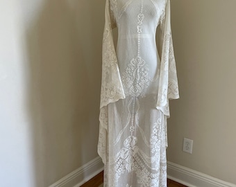 May include: Ivory lace dress with long, flared sleeves and intricate floral patterns. The dress is displayed on a mannequin, showcasing its delicate details and flowing silhouette. A vintage-inspired garment, perfect for special occasions.