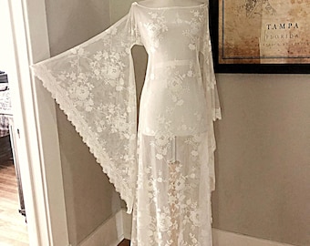 May include: A floor-length white lace dress featuring an off-the-shoulder neckline and wide, dramatic bell sleeves. The garment is crafted from a sheer, floral-patterned lace fabric that extends from the shoulders down to the hemline.