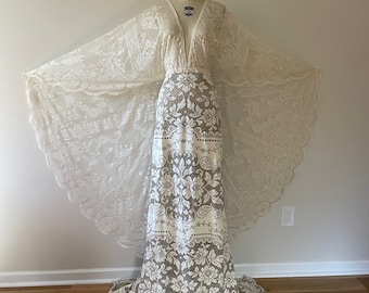 May include: A white lace wedding dress with a deep V-neckline and long, flowing sleeves. The dress has a fitted bodice and a flowing skirt with a delicate floral pattern.