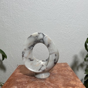 May include: A white marble sculpture featuring a smooth, circular, open-center design with prominent grey and dark brown veining throughout the stone. The piece is shaped like a continuous, twisted loop that rests on a small, circular pedestal base.