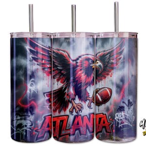 May include: Three stainless steel tumblers with lids and straws. Each tumbler features a design of a red and black eagle holding a football, with the word "ATLANTA" in red. The background has a graffiti-style design.