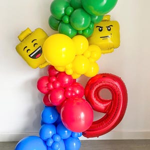 以下が含まれることがあります： 赤い数字の9、黄色のレゴ風バルーン、緑、赤、黄色、青のバルーンのクラスターが特徴的な、カラフルなバルーンアレンジメント。誕生日のお祝いに最適です。