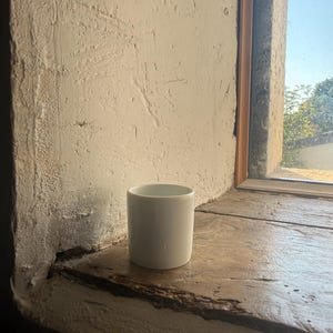 May include: A plain, cylindrical white ceramic mug with a smooth, glossy finish and a simple, handleless design. The mug features a clean, minimalist aesthetic with a uniform white colour throughout its exterior and interior surfaces.