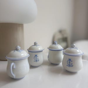 May include: Four small white porcelain lidded jars with blue trim and blue monogrammed lettering. Each jar features a rounded body, a matching lid with a small knob handle, and a delicate blue painted design on the exterior surface.