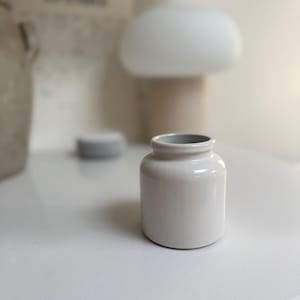 May include: A small, cylindrical white ceramic jar with a smooth, glossy finish and a short, slightly flared rim. The container features a simple, uniform design with a flat base and a wide opening, presenting a clean and neutral appearance.