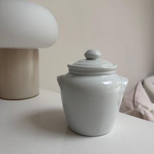 May include: A small, white ceramic jar with a lid. The jar has a rounded body with two small handles and a lid with a small knob. The jar is on a white surface. A beige lamp is in the background.