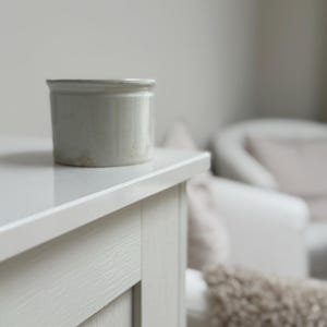 May include: A small, off-white ceramic jar with a slightly textured surface sits on a light-coloured table. The jar is cylindrical with a rounded rim. The table is part of a light-coloured cabinet with visible wood grain.