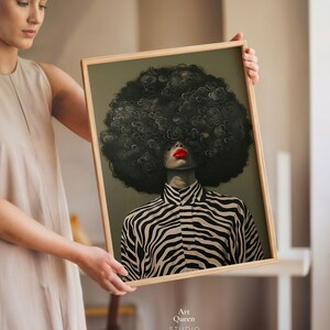 May include: A framed art print featuring a stylized portrait of a person with a large, dark, curly afro and red lipstick. The person is wearing a black and white zebra-striped shirt. The artwork is labeled "Art Queen Studio."