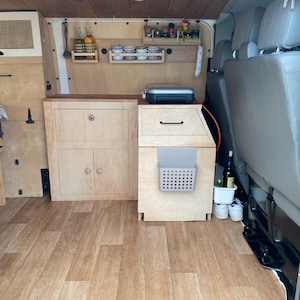 Puede incluir: El interior de una furgoneta camper con armarios de madera clara, techo de madera y suelo con aspecto de madera. La zona de cocina incluye una encimera, un hornillo portátil, estantes de pared con tazas y botellas, y un pequeño contenedor gris.