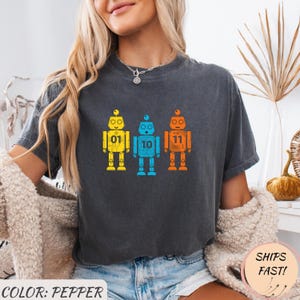 Może przedstawiać: Ciemnoszara koszulka z trzema kolorowymi ilustracjami robotów. Każdy robot jest w innym kolorze: żółtym, niebieskim i pomarańczowym, z numerami 01, 10 i 11 na piersi. Koszulka ma luźny krój.