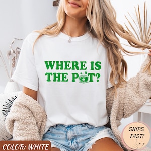 Puede incluir: Camiseta blanca con el texto verde "WHERE IS THE POT?" y un gráfico de una olla de oro y tréboles. La camiseta también tiene el texto "SHIPS FAST!" en un círculo rosa.