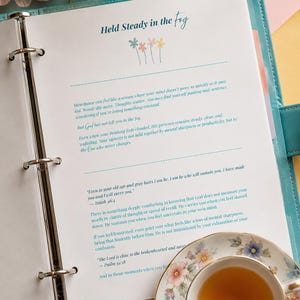 May include: A teal binder containing a page titled Held Steady in the fog with floral illustrations. The text discusses menopause and faith, including Bible verses from Isaiah 46:4 and Psalm 34:18. A floral teacup sits on the bottom right corner.