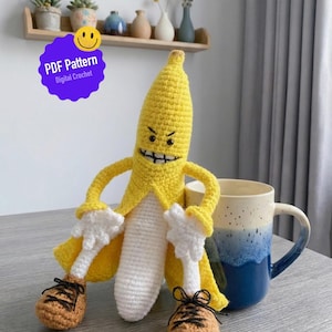 May include: A handmade crochet banana figure with a smiling face, arms, and legs. The banana is yellow with white gloves and a white body. It wears brown shoes with black laces. A blue and white mug sits nearby. The image includes a purple sticker that says "PDF Pattern Digital Crochet".