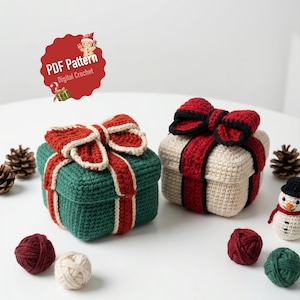 May include: Two crocheted gift boxes with red bows, one green and one beige, sit on a white surface. The green box has red and white ribbon accents. The image includes yarn balls and a snowman figurine. The text "PDF Pattern Digital Crochet" is visible.