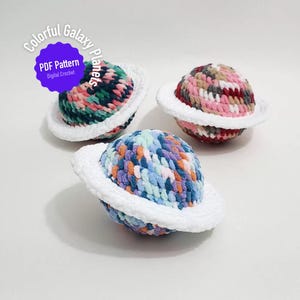 May include: Three crocheted planet toys in vibrant colours, each with a white ring. The planets are crafted from yarn in shades of pink, green, blue, and purple. The image includes text that reads "Colorful Galaxy Planets" and "PDF Pattern Digital Crochet".