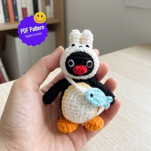 Może przedstawiać: Ręcznie robiony szydełkowy pingwin w białym kapeluszu z króliczymi uszami i jasnoniebieską torbą w kształcie ryby. Pingwin jest czarno-biały z pomarańczowymi stopami i czerwonym dziobem. Obraz zawiera tekst "PDF Pattern Digital Crochet".