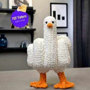May include: A handmade white crochet goose with orange feet and beak. The goose is standing upright. A purple badge with the text "PDF Pattern Digital Crochet" is in the upper left corner.