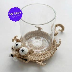 以下が含まれることがあります： 大きな白い目と歯を持つ漫画の生き物のような形をした、ベージュ色の手編みのコースターの上に、透明なガラスのコップが置かれています。コースターは質感のある外観です。「PDF Pattern Digital Crochet」というテキストの紫色のステッカーが画像にあります。