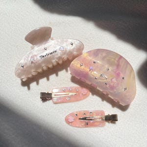 以下が含まれることがあります： 4つのヘアアクセサリーのセット。2つのヘアクリップと2つのヘアピン。ヘアクリップは淡いピンク色で、イリデッセントなアクセントと「Weritsere」の文字が入っています。ヘアピンも淡いピンク色で、花柄とラインストーンのディテールが施されています。