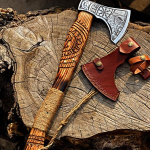 Könnte beinhalten: Eine handgefertigte Axt mit einem Holzgriff mit aufwendigen Schnitzereien und einer silbernen Klinge. Der Griff ist mit Bindfaden umwickelt, und eine braune Lederscheide ist enthalten. Die Axt wird auf einer Holzoberfläche präsentiert.